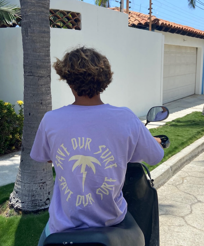 Person wearing a save our surf purple t-shirt with a palm tree design and text, sitting on a scooter.